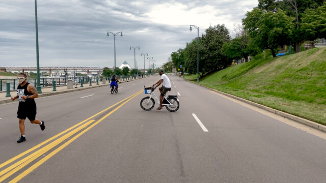 A man rides his bike down an open street as people run nearby.