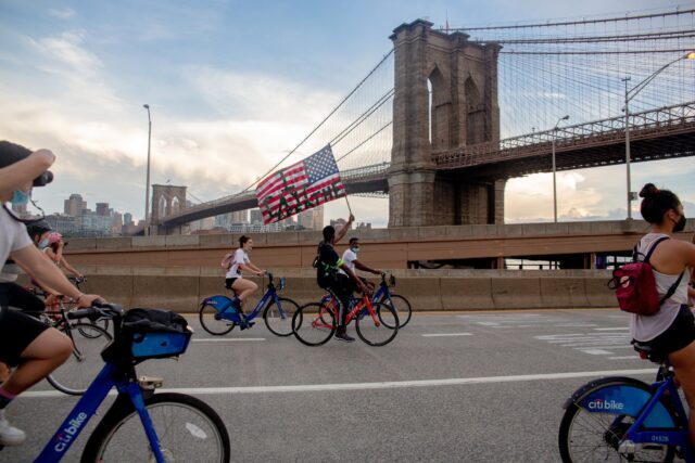 People on bikes ride across a bridge, and one person holds up an American Flag with the words "I can't breathe" written on it.